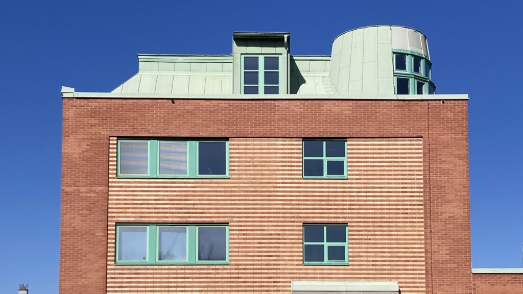 Tegelfasad på en byggnad i flera våningar med horisontellt mönstrat tegel och fönster med gröna karmar. Ovanför fasaden syns ett ljusgrönt plåttak med takkupor och ett runt hörnparti, mot en klar blå himmel.