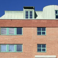 Tegelfasad på en byggnad i flera våningar med horisontellt mönstrat tegel och fönster med gröna karmar. Ovanför fasaden syns ett ljusgrönt plåttak med takkupor och ett runt hörnparti, mot en klar blå himmel.