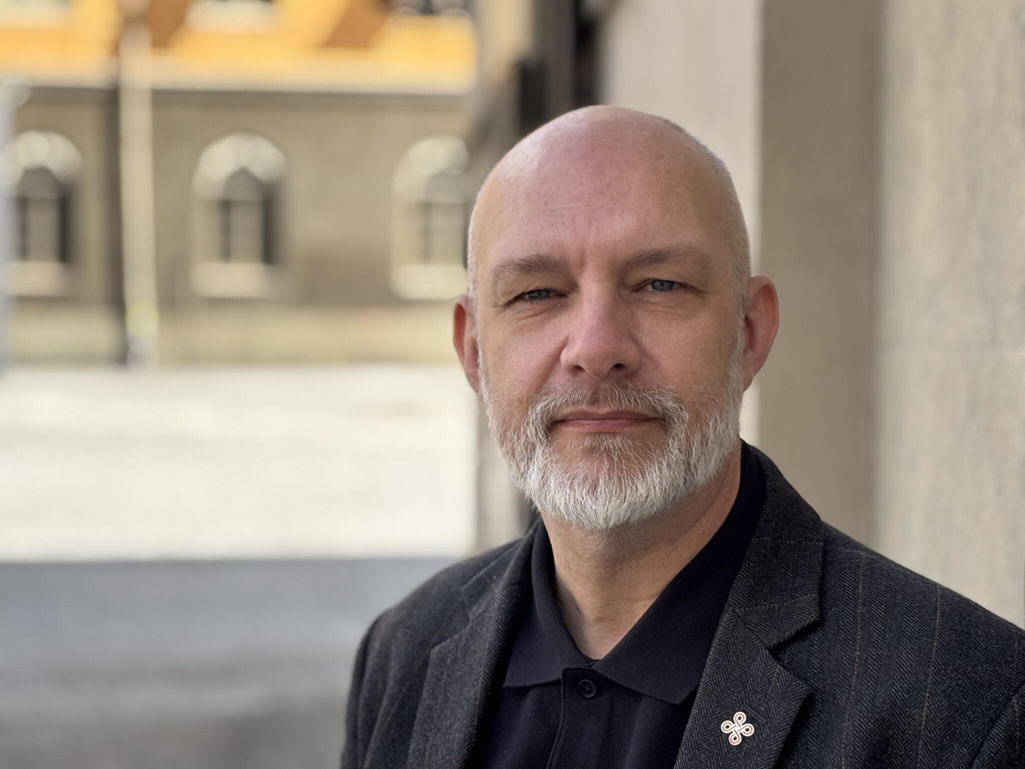 Vit man utan hår men med grått skägg står utomhus framför en grå fasad.