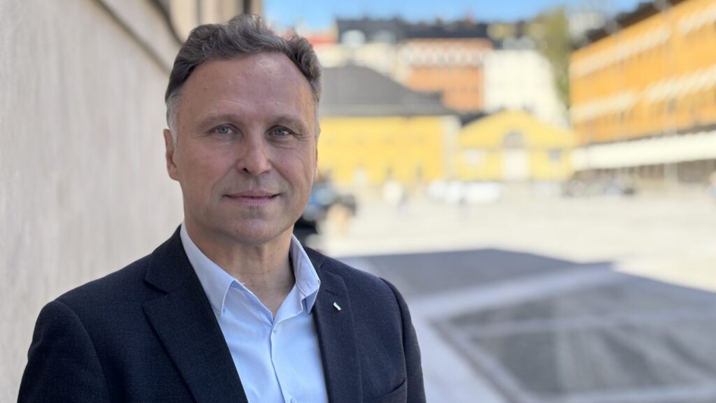Porträtt av en person i kavaj och ljus skjorta utomhus. I bakgrunden syns suddiga byggnader i stadsmiljö.