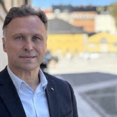 Porträtt av en person i kavaj och ljus skjorta utomhus. I bakgrunden syns suddiga byggnader i stadsmiljö.