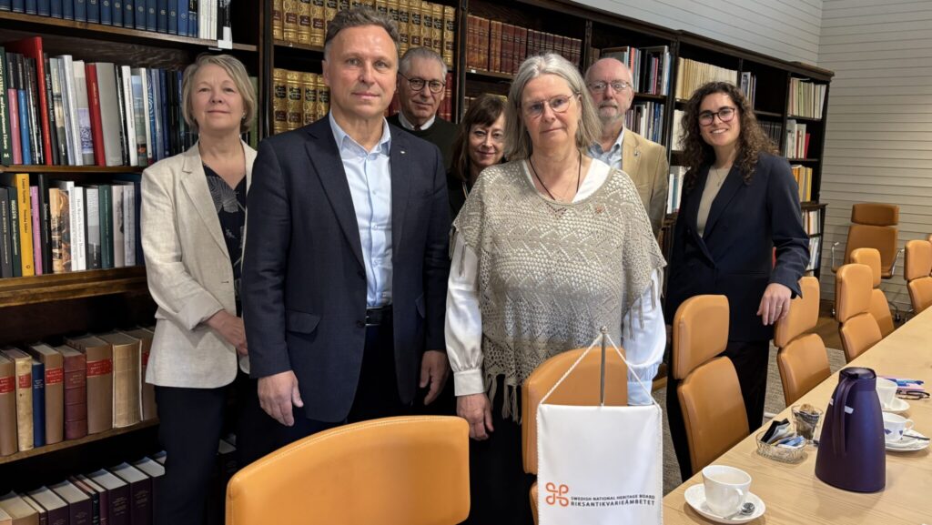 Gruppbild med åtta personer i ett biblioteksliknande mötesrum, med bokhyllor i bakgrunden. På ett konferensbord framför står ett standar med logo.