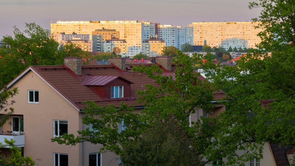 Bostadshus med ljus puts och röda tegeltak omgivna av grönskande träd i förgrunden, med höga flerbostadshus i ljusa färger i bakgrunden i kvällsljus.