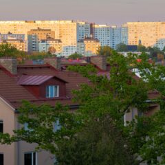 Bostadshus med ljus puts och röda tegeltak omgivna av grönskande träd i förgrunden, med höga flerbostadshus i ljusa färger i bakgrunden i kvällsljus.