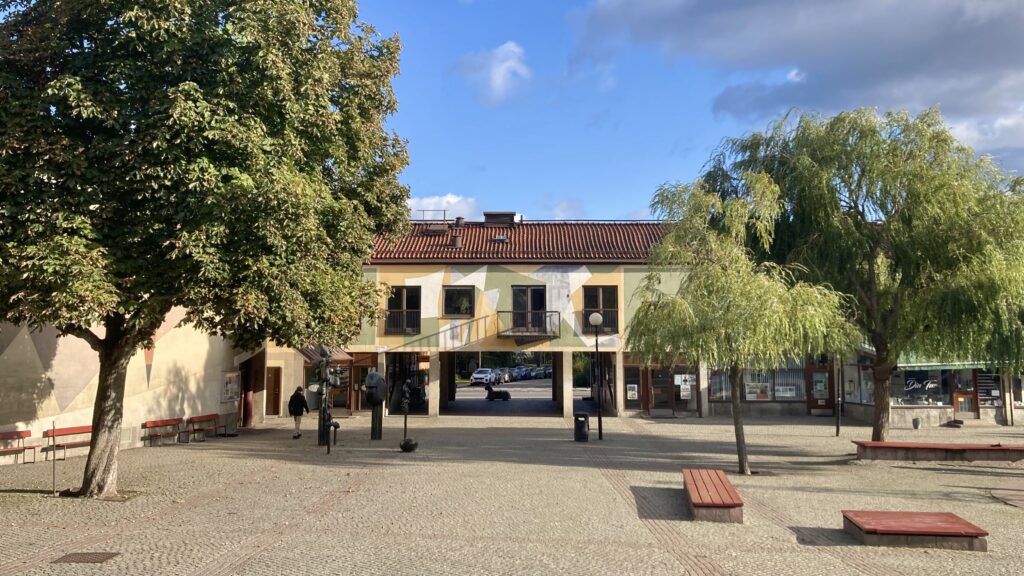 Torg med stenbelagd mark, bänkar och träd. I bakgrunden syns en låg byggnad med genomgång i bottenplan, balkonger på övre våningen och en fasad målad med geometriska mönster. Några personer rör sig över torget.