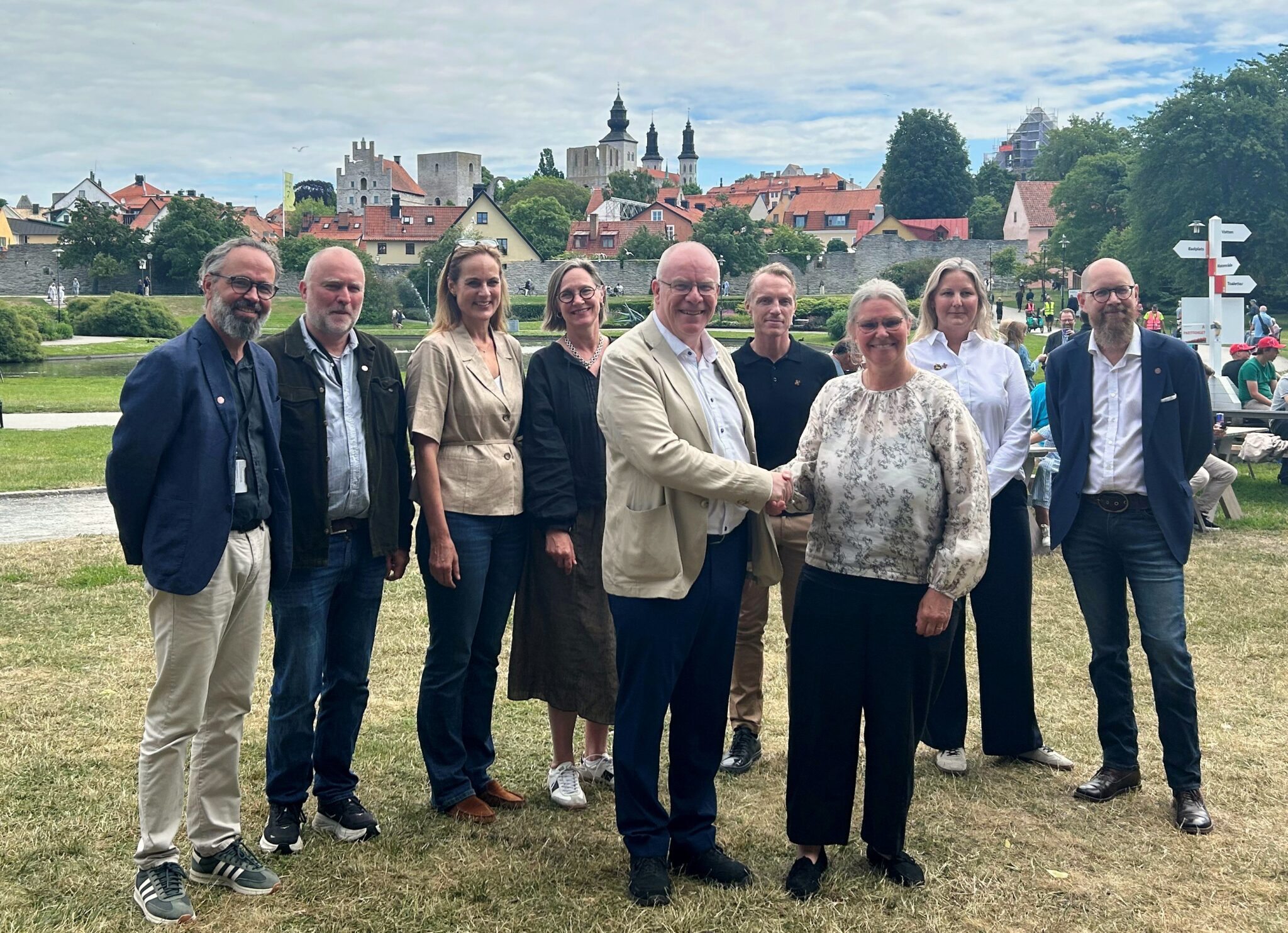 Grupp av tio personer som står utomhus i Almedalen i Visby, med ringmuren och kyrkspiror i bakgrunden; två personer i mitten skakar hand.
