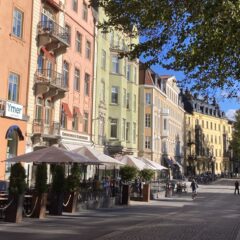 Färgglada hus längs solig stadsgata med uteservering och träd. Människor promenerar i bakgrunden.