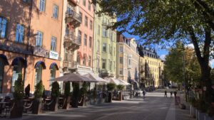 Färgglada hus längs solig stadsgata med uteservering och träd. Människor promenerar i bakgrunden.