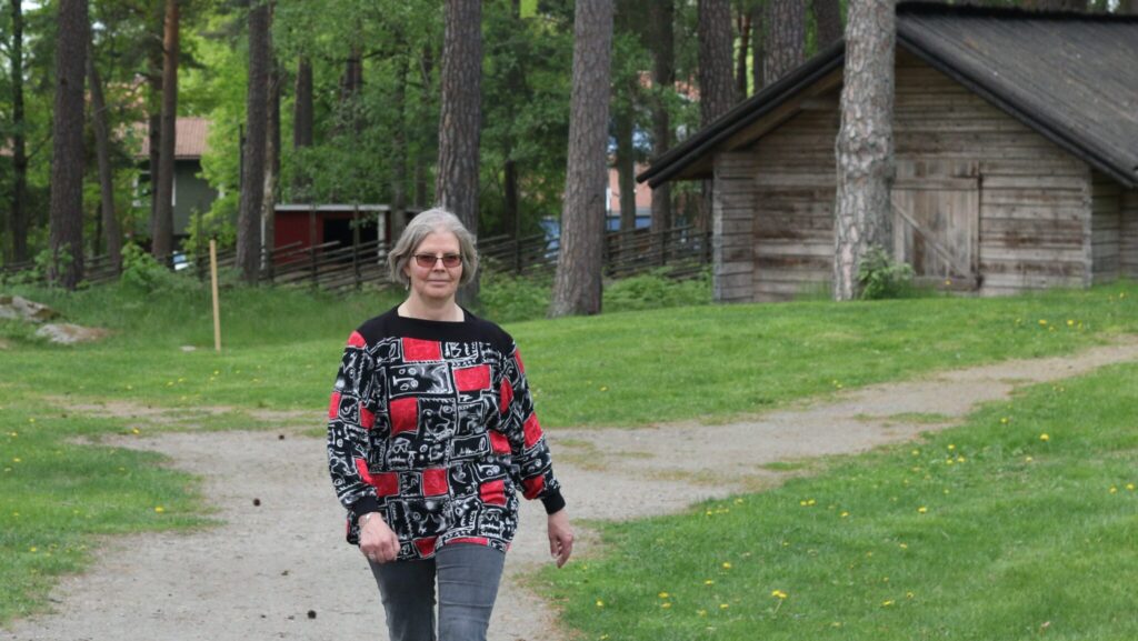 En kvinna i blommig tröja och jeans går på en grusgång mellan gräs och fäbod