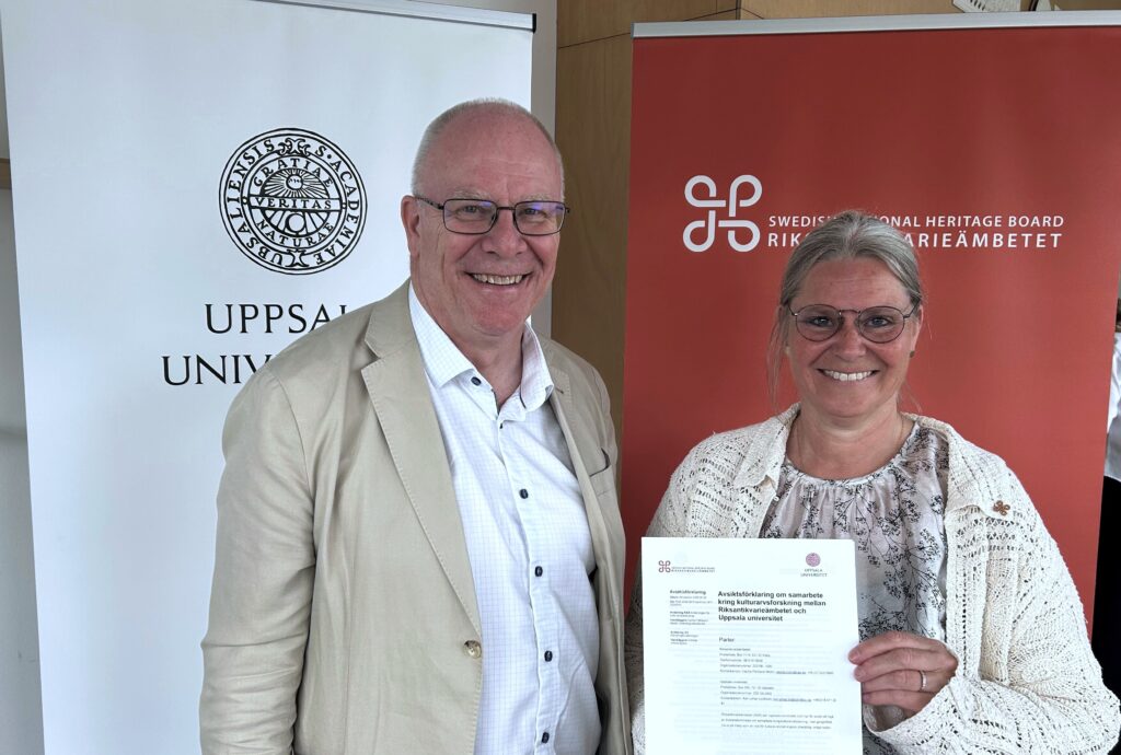 Två leende personer står framför rollups från Uppsala universitet och Riksantikvarieämbetet; den ena håller upp ett dokument om samarbete inom kulturarvsforskning.