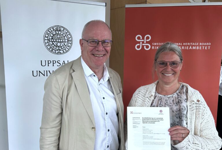 Två leende personer står framför rollups från Uppsala universitet och Riksantikvarieämbetet; den ena håller upp ett dokument om samarbete inom kulturarvsforskning.