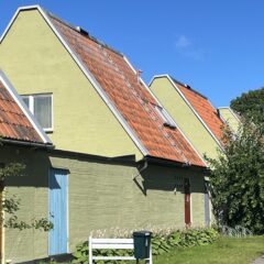Kedjehus med ljusgröna fasader och branta tegelklädda tak i rad, med små fönster, dörrar och grönska längs en gång under klarblå himmel.