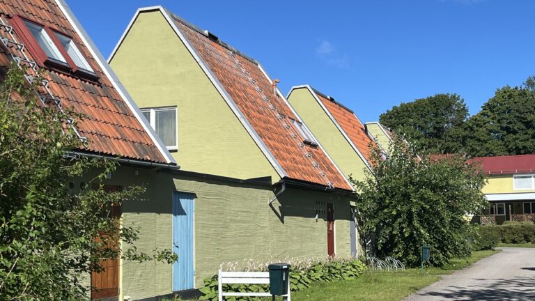 Kedjehus med ljusgröna fasader och branta tegelklädda tak i rad, med små fönster, dörrar och grönska längs en gång under klarblå himmel.