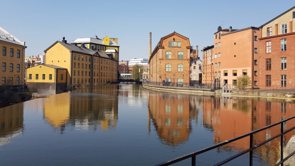 Färgstarka industribyggnader i tegel och gult fasadmaterial längs en stilla kanal, med speglingar i vattnet och en gångbro i bakgrunden.