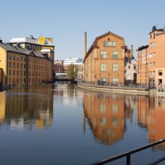 Färgstarka industribyggnader i tegel och gult fasadmaterial längs en stilla kanal, med speglingar i vattnet och en gångbro i bakgrunden.