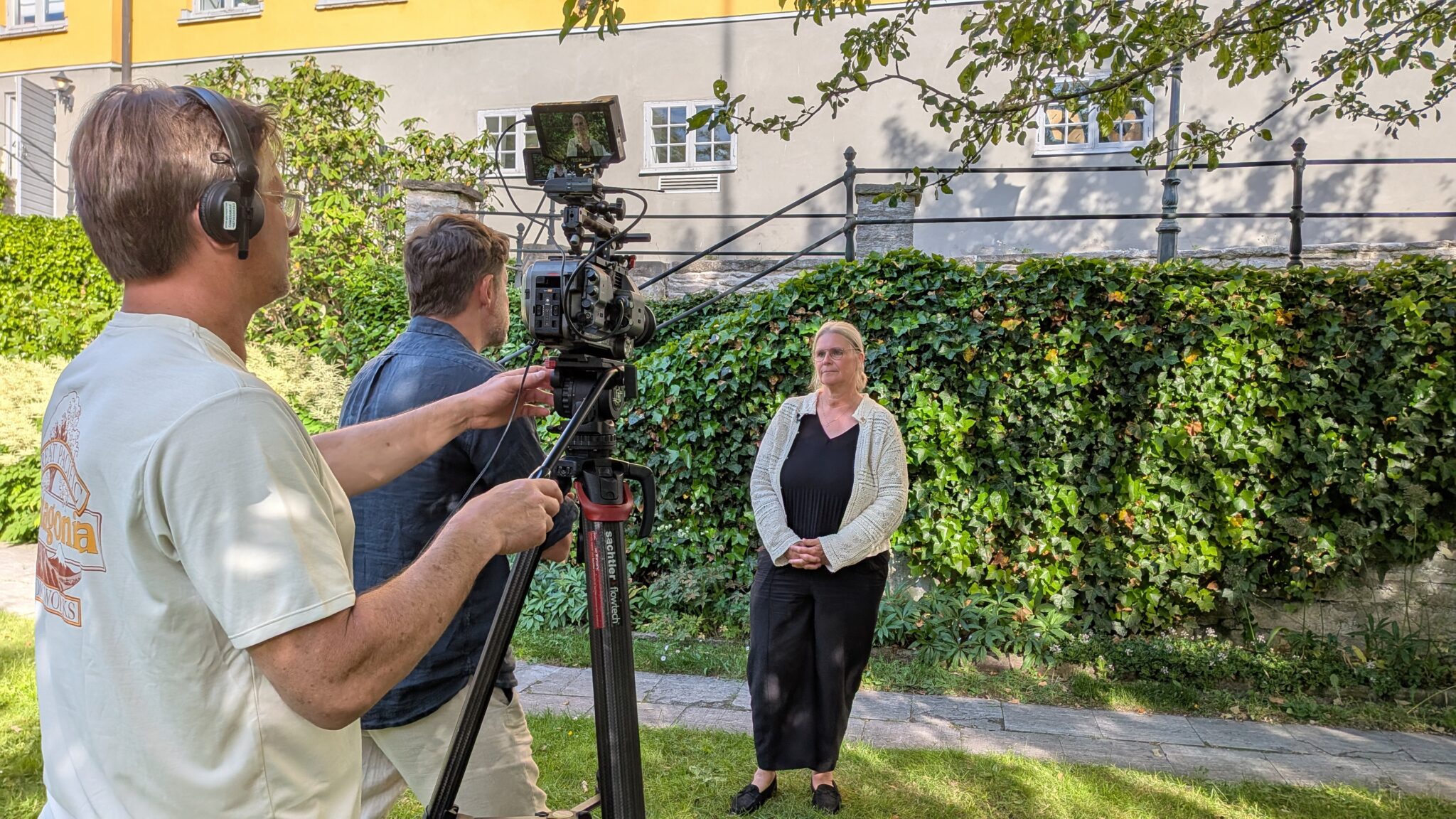 Filmteam spelar in en intervju utomhus; två personer står vid en kamera på stativ och filmar en person framför en mur med grönska och byggnader i bakgrunden.