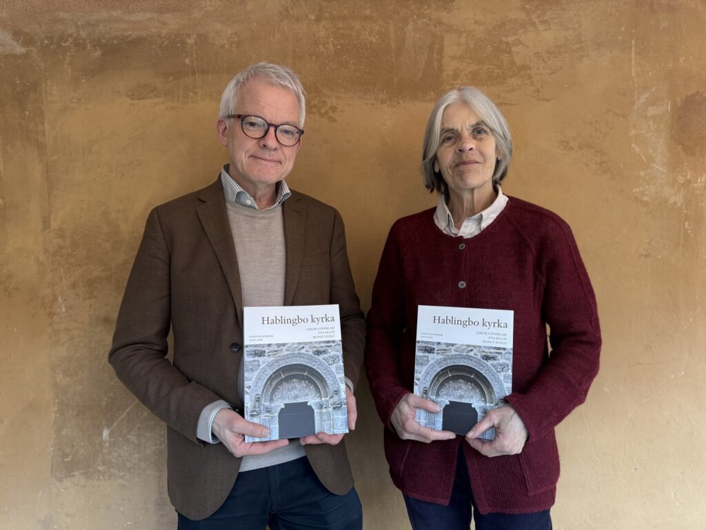 Halvifigursporträtt av Jakob och Eva som står mot en husvägg och håller ett varsitt exemplar av boken om Hablingbo kyrka