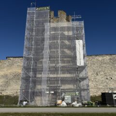 Ställning täcker ett högt sten­torn i en medeltida ringmur under renovering mot klarblå himmel.