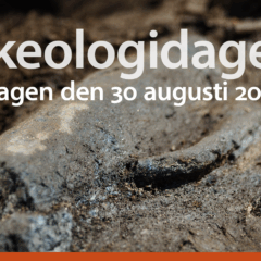Närbild på arkeologiskt fynd, en slags kanna som ligger halvt dold i jorden. På bilden ligger text med datum samt en symbol i orange.
