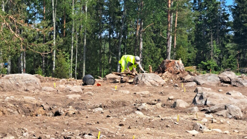 En arkeolog i gul jacka står böjd över marken och gräver i jorden i kanten av ett med pinnar utmärkt undersökningsområde