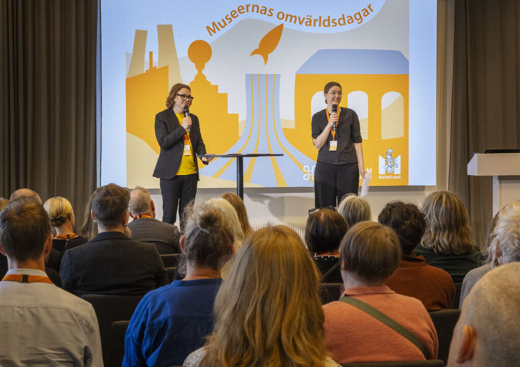 En man och en kvinna står på en scen och håller i mikrofoner inför en publik på en konferens.