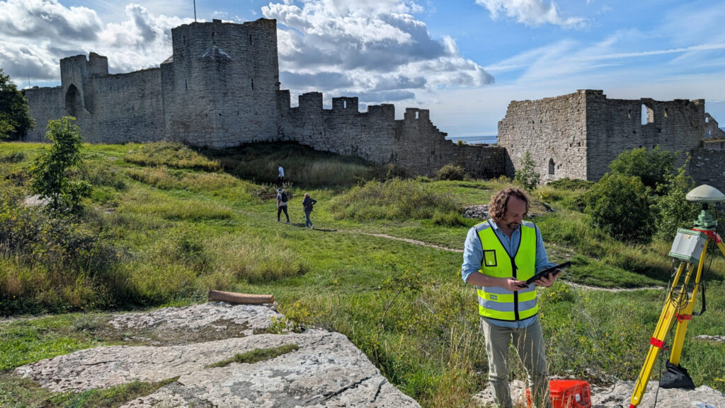 En man i gul varselväst står och granskar en datorskärm i handen. Bakom ser man ringmurens torn och däremellan en del utrustning som står och ligger på marken.