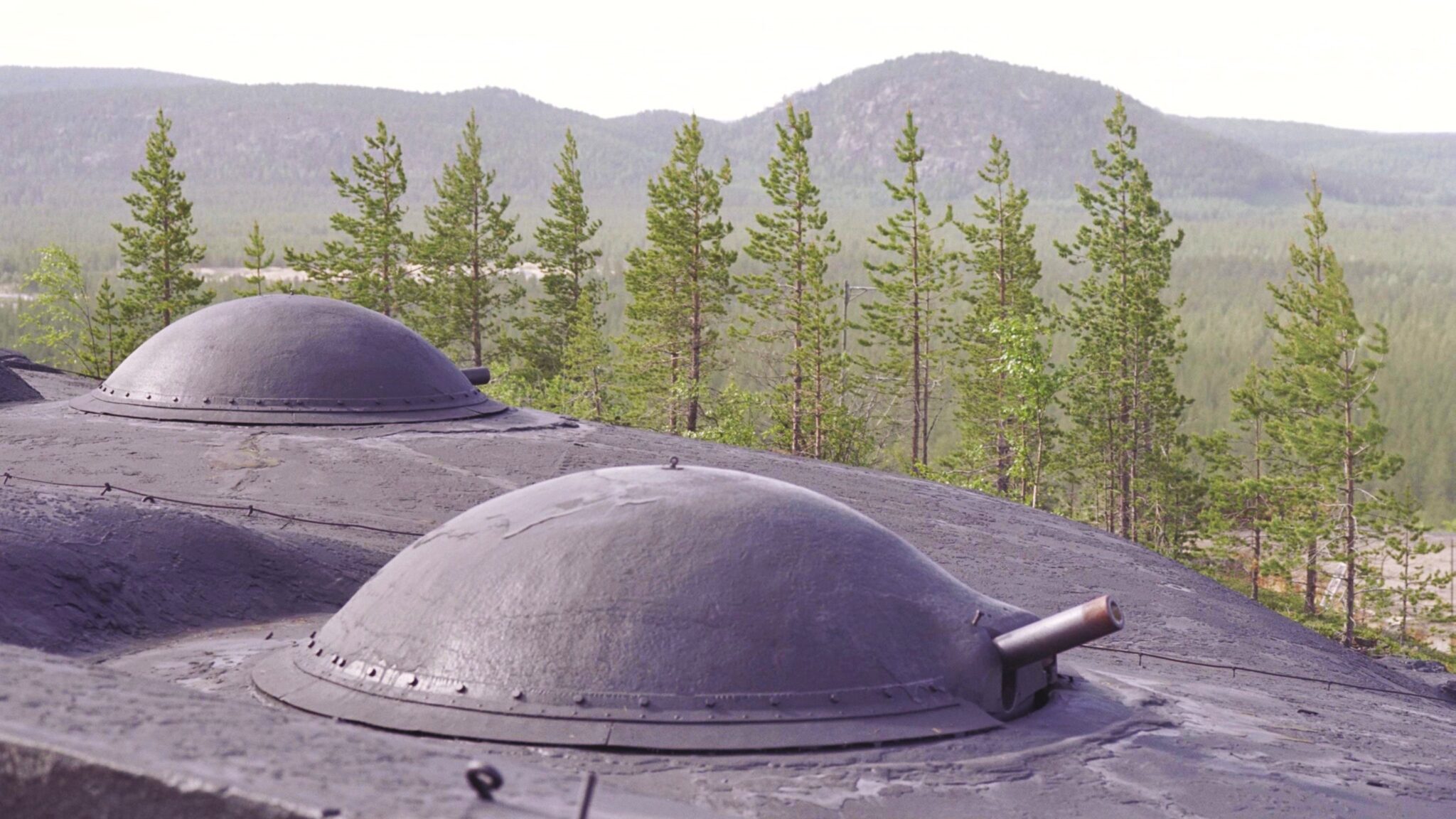 Två svarta försvarskupoler syns i fokus och i bakgrunden en rad granar. Bortom granarna syns höga berg.