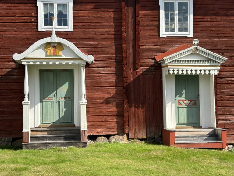 Närbild av fasaden på en traditionell hälsingegård i trä med två rikt dekorerade entréer, målade dubbeldörrar och snickarglädje