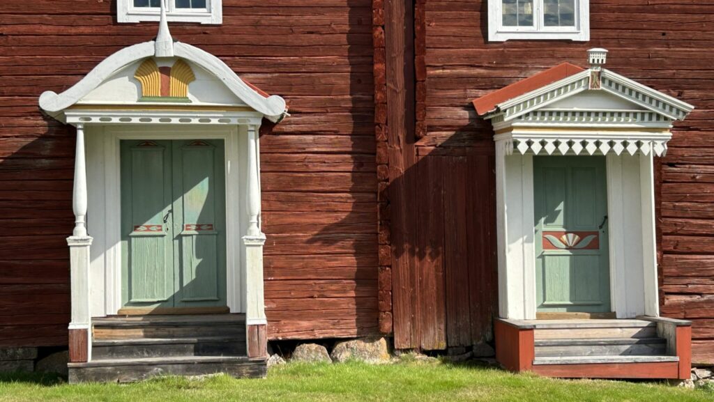 Närbild av fasaden på en traditionell hälsingegård i trä med två rikt dekorerade entréer, målade dubbeldörrar och snickarglädje