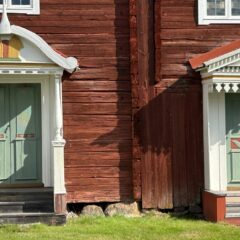 Närbild av fasaden på en traditionell hälsingegård i trä med två rikt dekorerade entréer, målade dubbeldörrar och snickarglädje