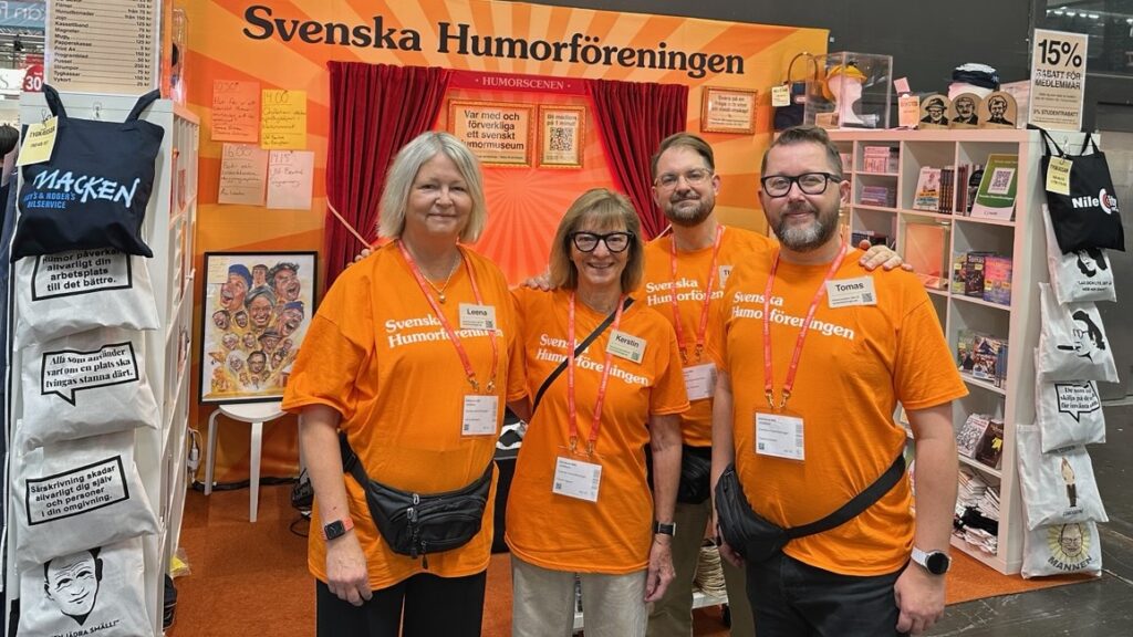 Två kvinnor och två män iklädda orange t-shirtar med texten "Svenska humorföreningen" i en monter.