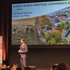 Två personer står på en scen framför en stor skärm med konferensbild från Visby och texten ”Nordic World Heritage Conference 2026”