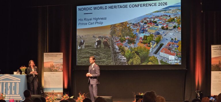 Två personer står på en scen framför en stor skärm med konferensbild från Visby och texten ”Nordic World Heritage Conference 2026”