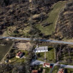 Flygfoto över en vit landsortskyrka med kyrkogård, omgiven av vägar, åkrar och gles lövskog i ett kuperat landskap.