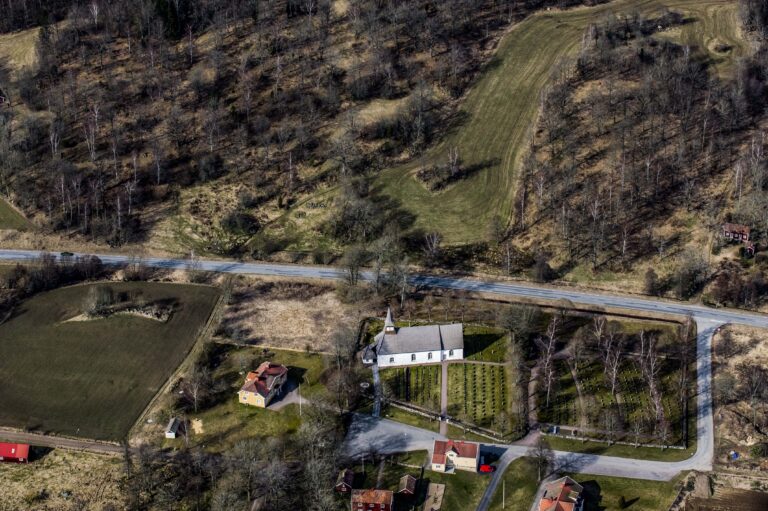 Flygfoto över en vit landsortskyrka med kyrkogård, omgiven av vägar, åkrar och gles lövskog i ett kuperat landskap.