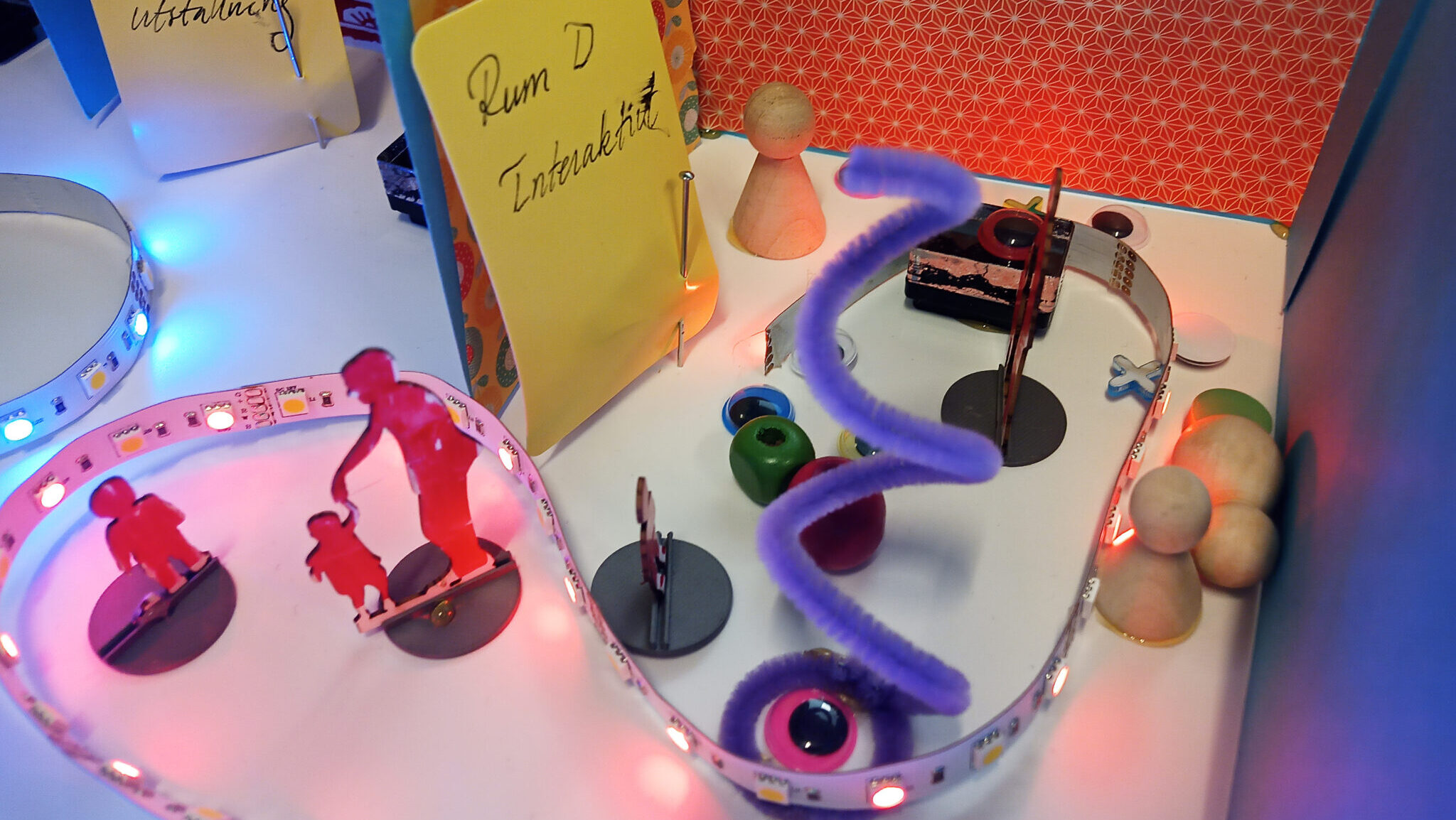 Modell av en interaktiv utställning med färgade ljusslingor, figurer, träklossar och en lila spiral. Skyltar markerar rum för interaktivitet och tillfällig utställning.