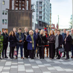 Gruppfoto med ett femtontal personer som står på ett torg mellan moderna byggnader. I gruppen syns personer i civila kläder, en person i militäruniform och en person i samisk kolt. I bakgrunden syns stadsmiljö och flerbostadshus.