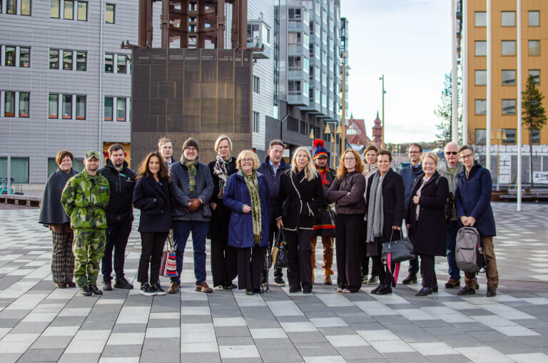 Gruppfoto med ett femtontal personer som står på ett torg mellan moderna byggnader. I gruppen syns personer i civila kläder, en person i militäruniform och en person i samisk kolt. I bakgrunden syns stadsmiljö och flerbostadshus.