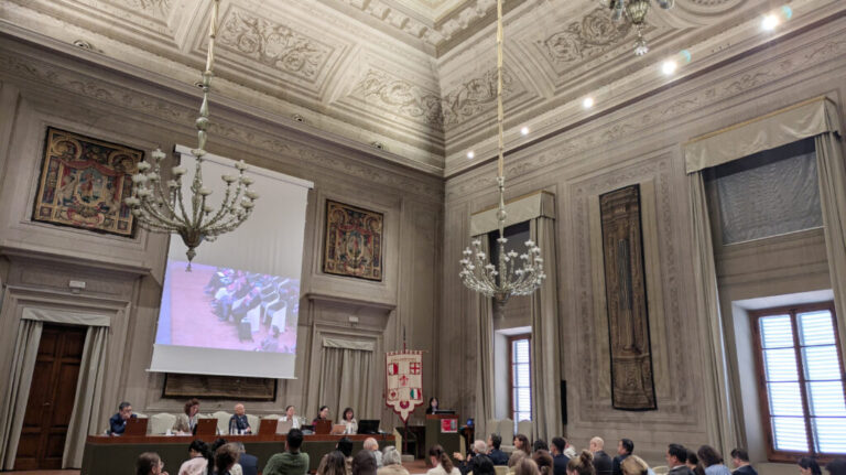 Stor konferenssal med högt dekorerat tak och flera stora kristallkronor. Längst fram sitter en panel vid ett bord framför en projektorduk som visar en presentation. Publik sitter på stolar riktade mot scenen. Rummet har höga fönster, väggdekorationer och en fana placerad bredvid talarstolen.