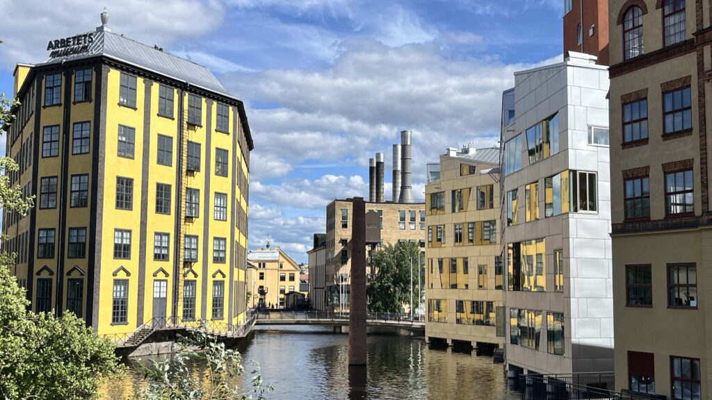 Gul industribyggnad och moderna hus speglas i en kanal under blå himmel med moln.