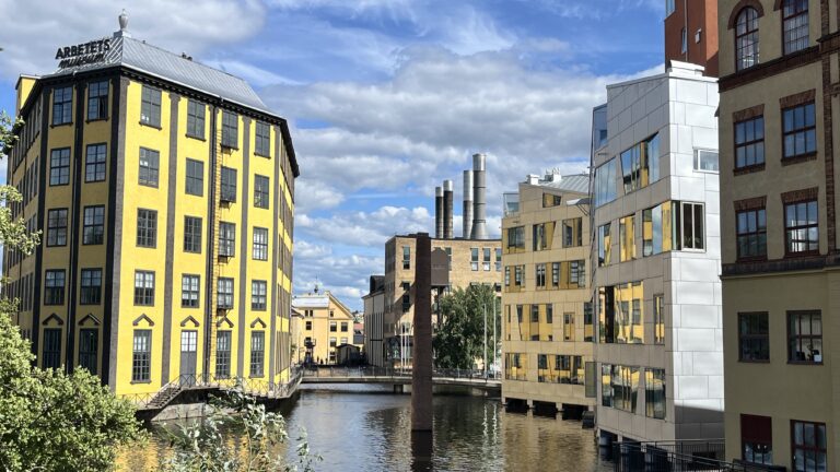 Gul industribyggnad och moderna hus speglas i en kanal under blå himmel med moln.