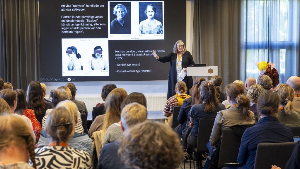 En kvinna står vid en pulpet och föreläser inför publik, en projektor visar fotografier från tidigt 1900-tal föreställande "rastyper".