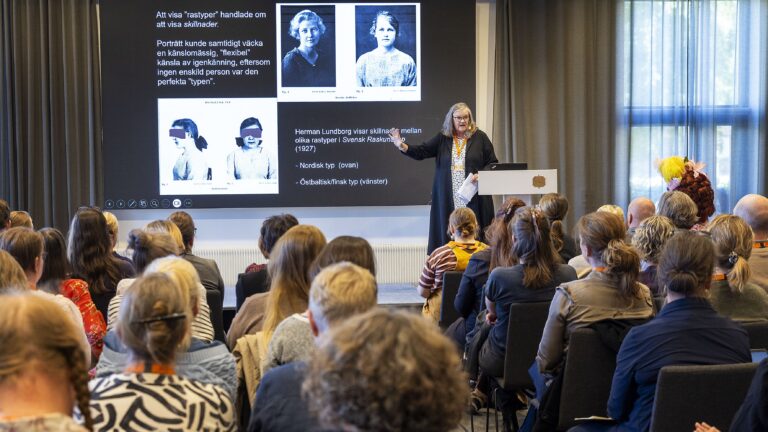 En kvinna står vid en pulpet och föreläser inför publik, en projektor visar fotografier från tidigt 1900-tal föreställande "rastyper".