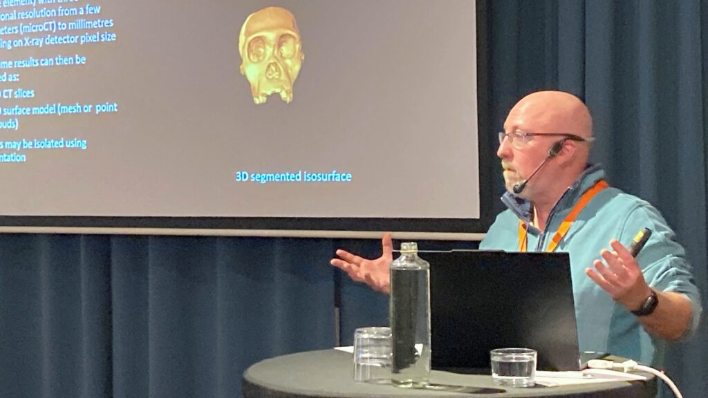 Person med headset föreläser vid ett bord med dator. På presentationsskärmen visas en 3D-bild av en skalle och text om digital bildanalys och CT-teknik.