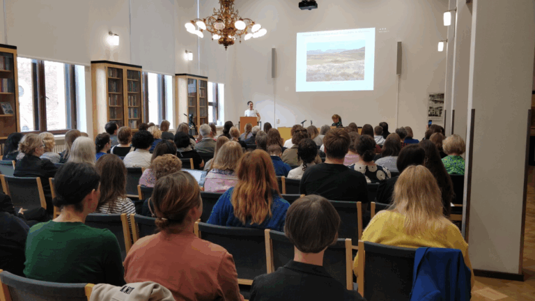 En konferenssal fylld med åhörare, en person står i talarstolen medan en projektion visas.