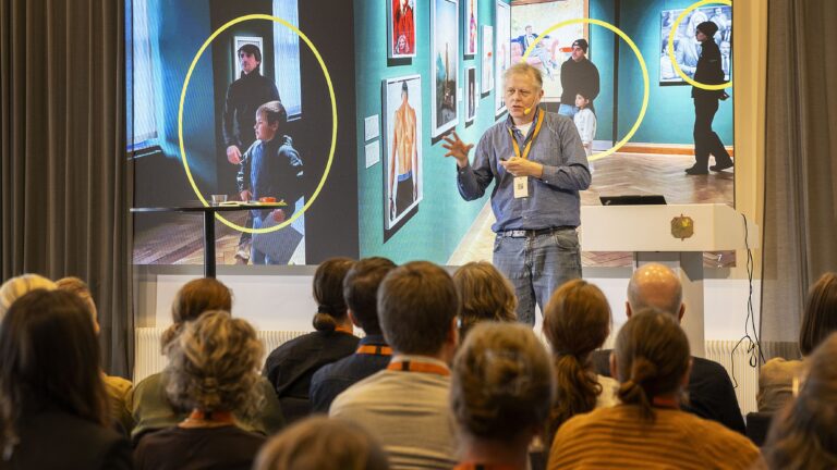 En man föreläser för en publik, i bakgrunden syns ett fotografi med gula cirklar från ett museum.