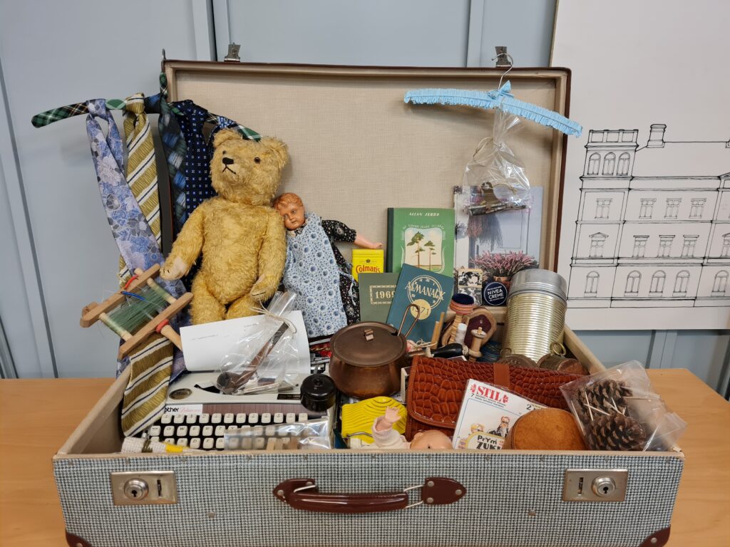 Bilden visar en öppen väska full med saker som en teddybjörn, skrivmaskin, termos, böcker och en docka.