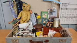 Bilden visar en öppen resväska av äldre modell full med saker som en teddybjörn, skrivmaskin, termos, böcker och en docka.